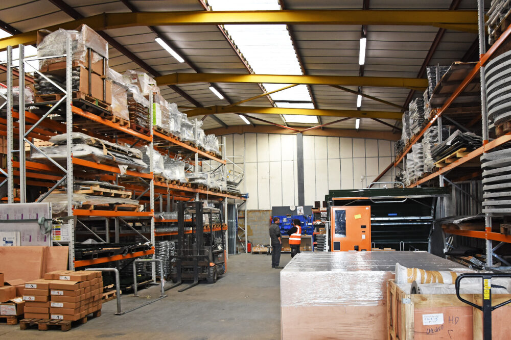 Two men stand talking inside a large warehouse with shelves of boxes and forklifts.
