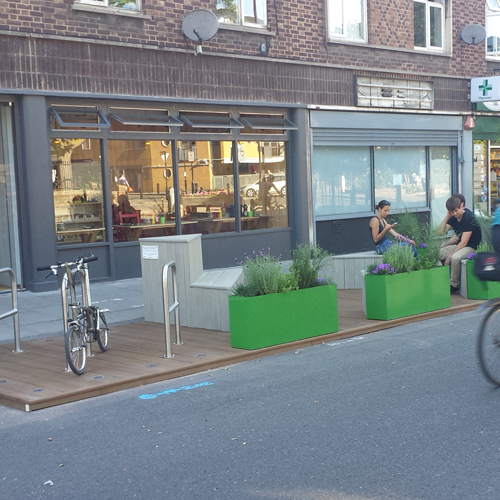 A street with a car space converted into a 'parklet' with seating and planters, in which two people are sitting.