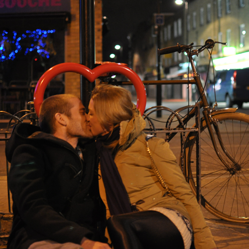 A couple kiss at night in front of a heart-shaped metal Lovehoop bike stand attached to a street post.