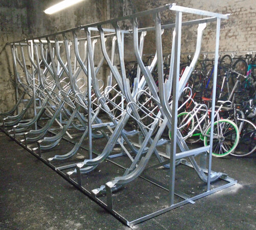 An empty metal rack for vertical bike storage, with a full rack behind it, both inside a concreted room.