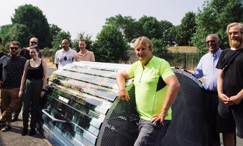 A group of smiling people stand around a dome-roofed Bikehangar bike storage unit on a leafy street.