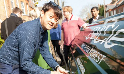 A man looks towards the camera while unlocking the door of a Bikehangar bike storage container on a residential street, with a group of people talking amongst themselves in the background.
