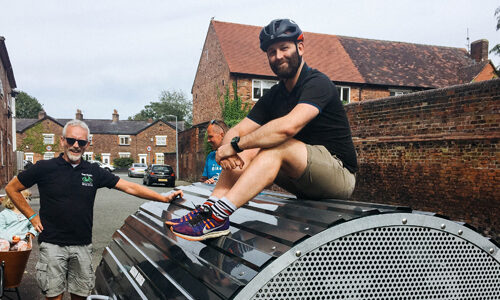 A man in a bike helmet sits on the domed roof of a metal Bikehangar next to a man who is leaning on the storage unit with his arm outstretched, on a residential street.