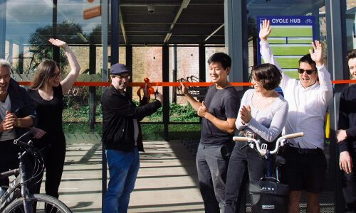 A group of people with bikes clap and celebrate as one man cuts a red ribbon surrounding a glass-walled Cycle Hub.